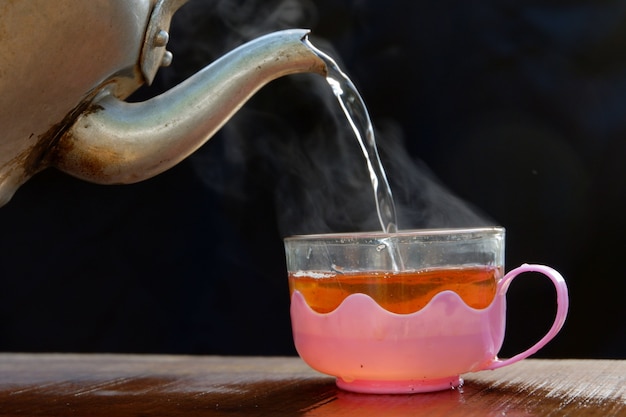 La Tasse De Thé Chaud A De La Vapeur Chaude Avec Une Bouilloire. Placé Sur Une Vieille Table En Bois, Fond Noir