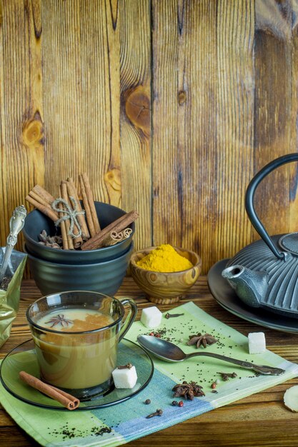 Une tasse de thé masala indien traditionnel avec des ingrédients à préparer. Cannelle, anis, sucre, thé noir, poivre, clous de girofle, curcuma sur une table en bois.