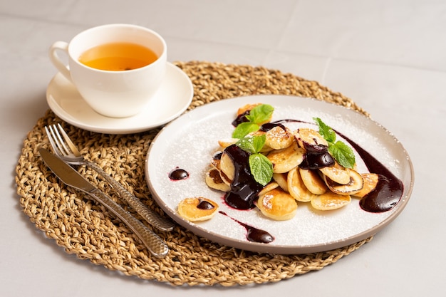 Une tasse de thé et des mini-crêpes avec de la confiture de petits fruits et de la menthe saupoudrée de sucre en poudre.