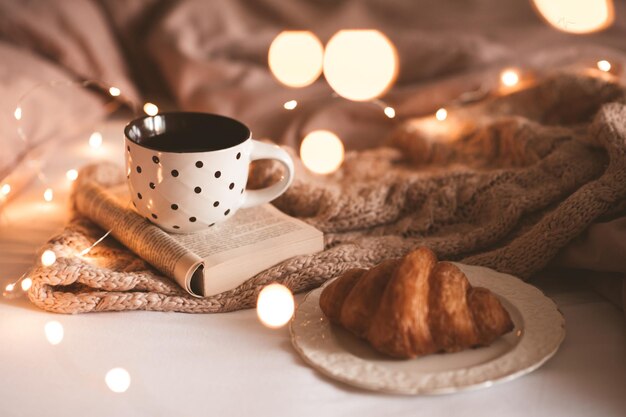 Tasse de thé noir frais sur un livre ouvert avec un délicieux croissant et du textile tricoté au lit sur des lumières rougeoyantes. Saison des vacances d'hiver.