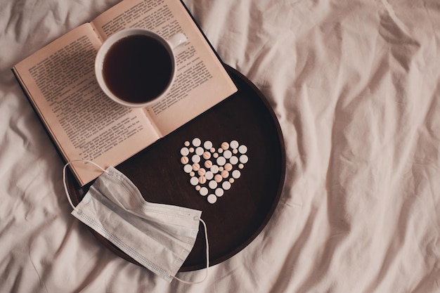 Tasse de thé restant sur un livre ouvert avec des médicaments en forme de coeur et un masque médical sur un plateau en bois au lit en gros plan. Bonjour. Quarantaine à la maison. Notion de virus.