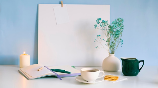 Une tasse de thé se dresse sur une table blanche avec un cahier, un stylo, une bougie allumée. Préparation à la rédaction du texte.