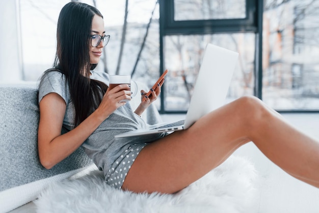 Avec une tasse de thé et un smartphone. Sexy belle jeune femme allongée sur le lit avec un ordinateur portable.