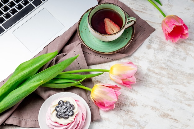 Une tasse de thé, des tulipes et un gâteau à côté d'un ordinateur portable sur la table.