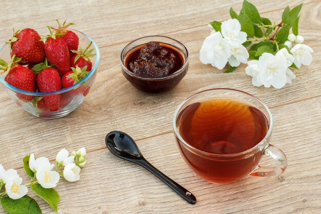 Tasse de thé en verre, confiture de fraises maison et fraises fraîches dans des bols, cuillère et fleurs de jasmin blanc sur des planches en bois. Vue de dessus.