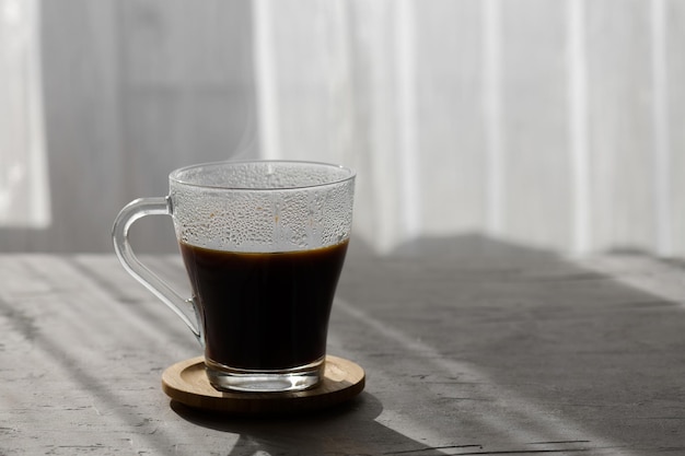 Une tasse transparente de café noir infusé chaud avec des ombres du matin et des rayons de soleil à travers la fenêtre sur fond de table gris