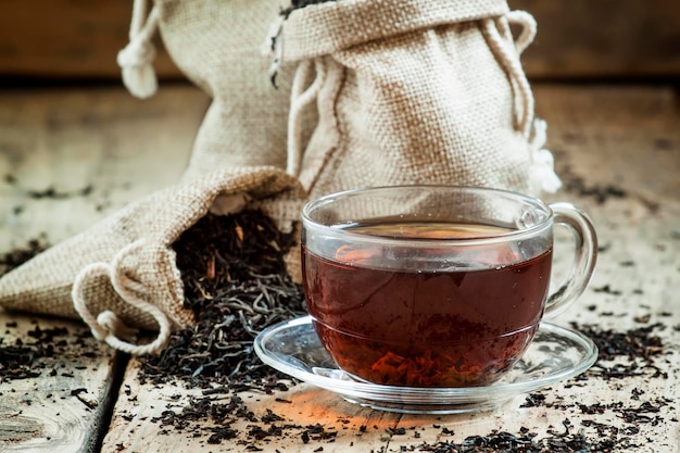 Tasse transparente en verre de thé noir et de thé sec dans des sacs sur le fond des feuilles de thé éparpillées sur le vieux fond en bois mise au point sélective