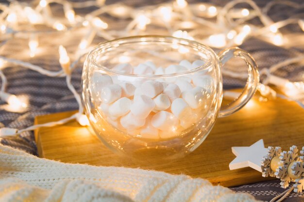 Tasse en verre avec guimauves sur planche de bois avec lumières de Noël et plaid doux. Flou artistique