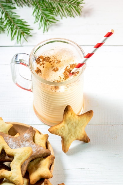 Tasse en verre avec lait de poule de Noël avec des biscuits en forme d'étoiles sur fond de bois blanc.