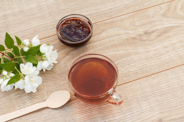 Tasse en verre de thé vert, confiture de fraises maison dans un bol en verre et fleurs de jasmin blanc sur fond de bois. Vue de dessus avec copie espacée.