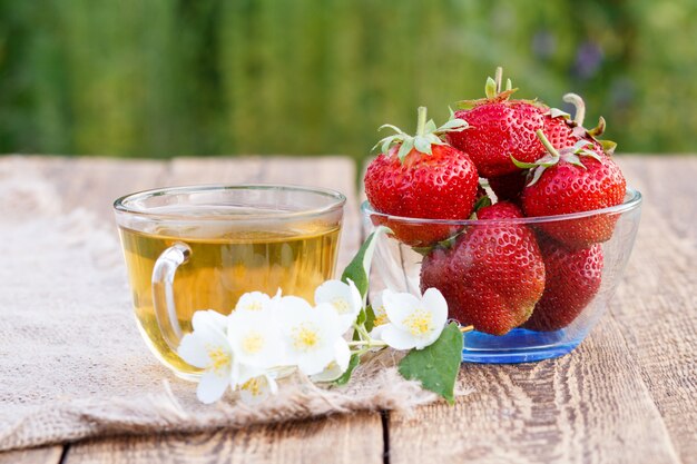 Tasse en verre de thé vert et de fraises mûres rouges dans un bol en verre avec des fleurs de jasmin sur un sac et de vieilles planches de bois avec un arrière-plan naturel flou