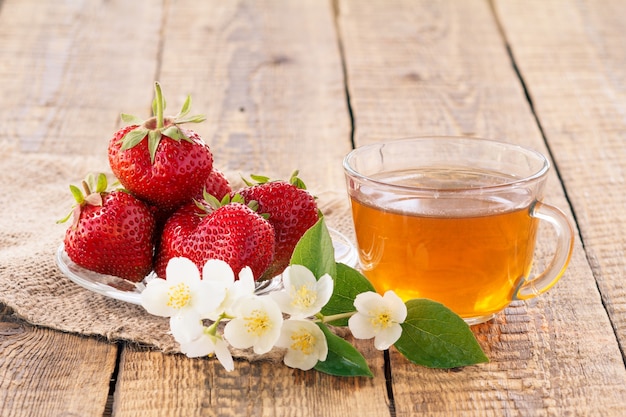Photo tasse en verre de thé vert et de fraises mûres rouges sur une soucoupe en verre avec des fleurs de jasmin sur un sac.