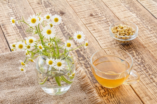 Tasse en verre de thé vert, pot avec des fleurs de camomille blanches sur un sac et petit bol en verre avec des fleurs sèches de matricaria chamomilla sur fond de bois