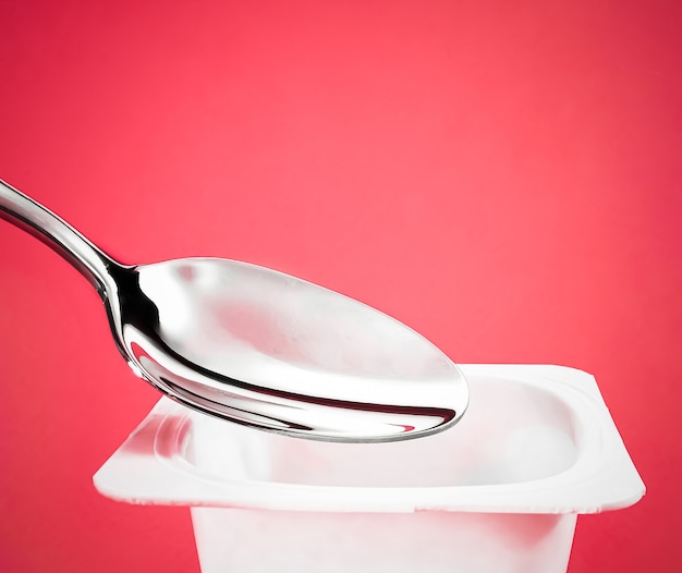 Tasse de yaourt et cuillère en argent sur fond rouge contenant en plastique blanc avec crème de yaourt produit laitier frais pour une alimentation saine et un équilibre nutritionnel