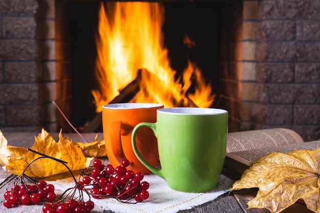 Tasses de thé ou de café, feuilles d'automne et baies de viorne devant une cheminée confortable dans une maison de campagne aut...