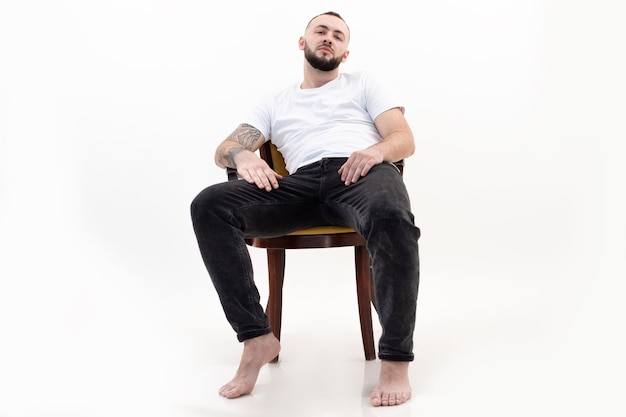 Tatoué beau brunet barbu barbu aux pieds nus assis relaxant comme le patron sur une chaise comme le patron