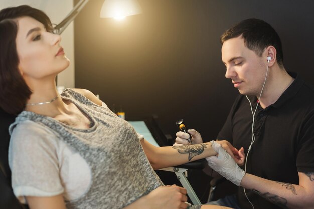 Tatoueur concentré travaillant avec une aiguille sur le bras des clients et écoutant de la musique dans des écouteurs, faisant abstraction de tout, de l'espace de copie