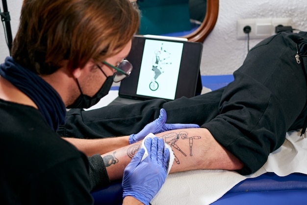 Un tatoueur en gants bleus regardant une tablette et faisant un tatouage sur la jambe d'un homme dans un studio