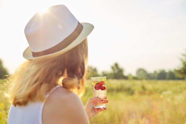 Été, chaleur, soif. Femme au chapeau avec son dos avec un verre de boisson rafraîchissante à la menthe aux fraises naturelles faites maison, espace de copie été coucher de soleil prairie nature