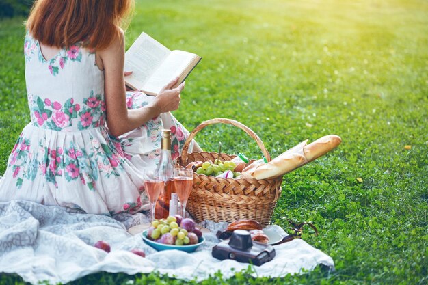 Été - pique-nique dans le pré. fille assise lisant un livre et près d'un panier de pique-nique et d'une baguette, du vin, des verres, des raisins et des petits pains