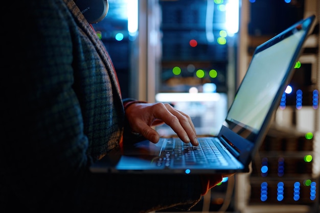 Technicien informatique vérifiant les éléments vitaux des serveurs à l'aide d'un ordinateur portable. Agent de soutien masculin travaillant dans une salle de serveur de réseau sombre