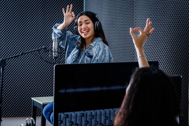 Technicien de studio féminin de l'écran d'ordinateur du logiciel pour l'enregistrement au premier plan d'une jeune chanteuse asiatique portant des écouteurs enregistrant une chanson devant un microphone dans un studio professionnel