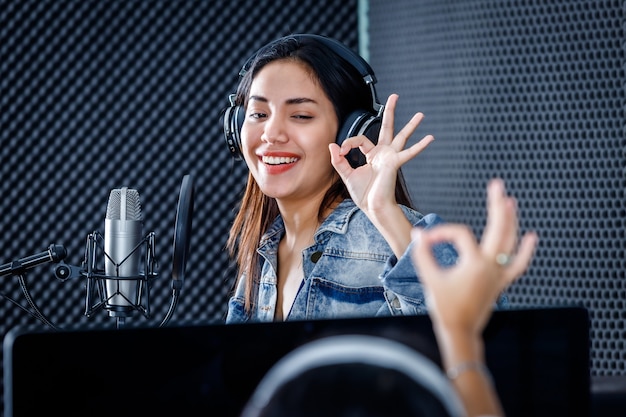 Technicien de studio féminin de l'écran d'ordinateur du logiciel pour l'enregistrement au premier plan d'une jeune chanteuse asiatique portant des écouteurs enregistrant une chanson devant un microphone dans un studio professionnel