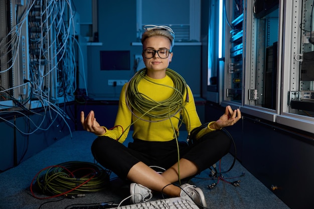 Technicienne informatique calme enveloppée dans des fils Internet et des câbles de réseau méditant alors qu'elle est assise en position de lotus sur le sol d'une salle de serveur avec un rack de superordinateur