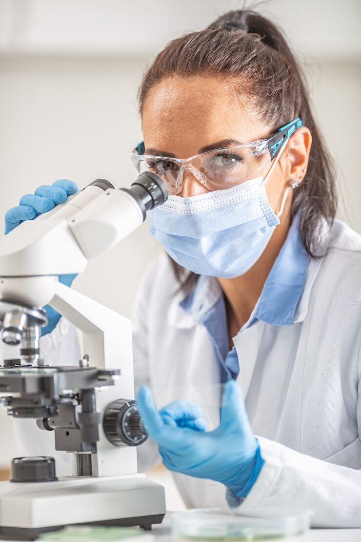 Photo une technicienne de laboratoire portant des lunettes de protection, des gants et un masque facial est assise à côté d'un microscope et d'une fiole conique, regardant dans la caméra.