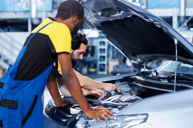 Photo techniciens collaborant à l'entretien d'un véhicule cassé, vérification de la courroie de compresseur défectueuse, réparateurs compétents dans un atelier de réparation automobile travaillant ensemble à la réparation d'une automobile, discutant des meilleures options.