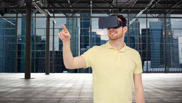 Technologie 3d, réalité virtuelle, concept de divertissement et de personnes - jeune homme heureux avec un casque de réalité virtuelle ou des lunettes 3d jouant à un jeu sur une salle vide industrielle et un arrière-plan panoramique de la ville