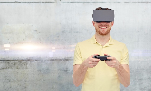 Technologie 3d, réalité virtuelle, concept de divertissement et de personnes - jeune homme heureux avec un casque de réalité virtuelle ou des lunettes 3d jouant avec une manette de jeu sur fond de mur en béton gris
