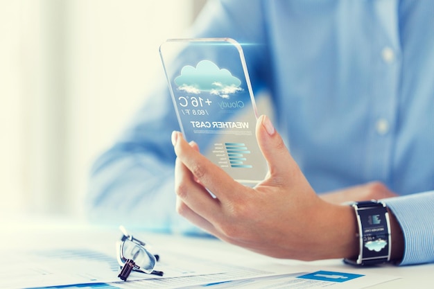 technologie, application, météo, prévisions et concept de personnes - gros plan sur une main de femme tenant un smartphone transparent avec icône nuage et température de l'air à l'écran au bureau