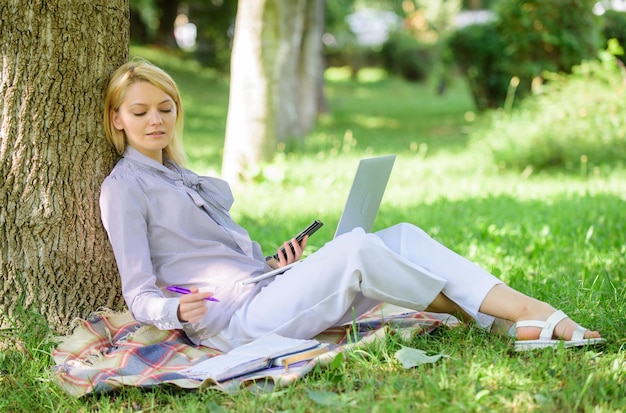 Technologie de l'éducation et concept internet. Une femme avec un ordinateur portable travaille à l'extérieur s'appuie sur un tronc d'arbre. Fille travaille avec un ordinateur portable dans le parc assis sur l'herbe. Bureau de l'environnement naturel. Avantages du travail à l'extérieur.