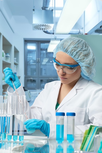 La technologie féminine pharmaceutique travaille en laboratoire. Jeune femme caucasienne portant des gants de protection, un chapeau et une robe blanche, échantillon de pipettes en flacon. Intérieur du laboratoire, mise au point sélective. Recherche en biotechnologie.