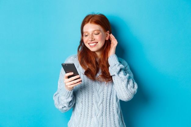 La technologie. Fille rousse heureuse regardant heureux au téléphone mobile, lisant le compliment dans le message, souriant et rentrer les cheveux derrière l'oreille, debout en pull sur fond bleu.