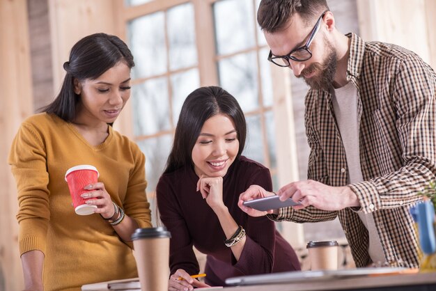 Technologie moderne. Agréable bel homme barbu tenant son smartphone et prenant une photo en se tenant debout avec ses collègues autour de la table