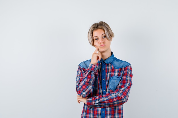 Teen boy holding doigt sur la joue en chemise à carreaux et à la pensive , vue de face.