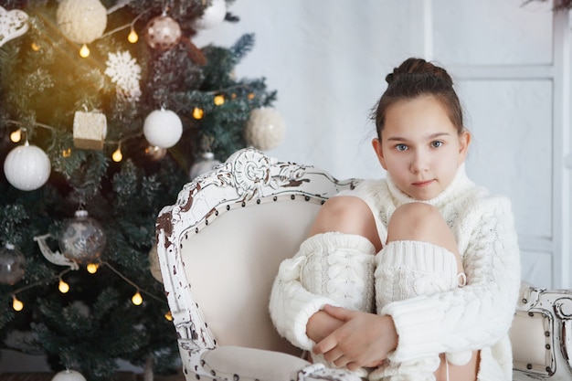 Teen girl assis sur une chaise près de l'arbre de Noël. Vêtu d'un pull en laine tricoté blanc et de leggings. Les cheveux sont groupés. Une agréable surprise. Cadeaux du nouvel an.