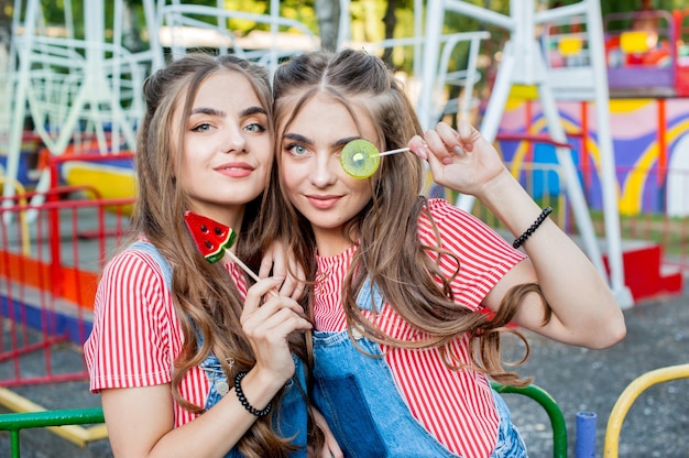 Teen jumelles dans des vêtements colorés avec des sucettes au caramel avec des attractions foraines colorées