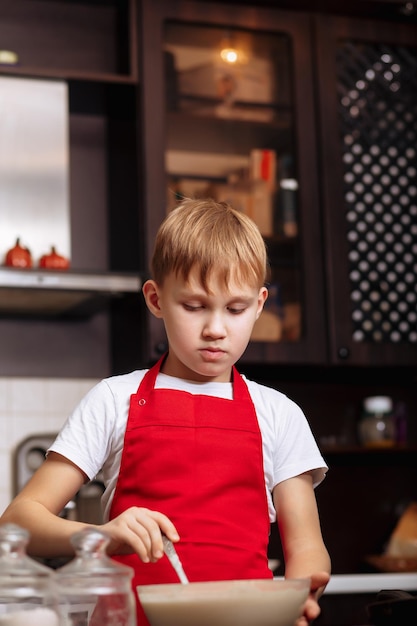 Teenage Boy en tablier rouge dans la cuisine à la maison faire de la pâte pour la pâtisserie de crêpes Concept de cuisine pour enfants