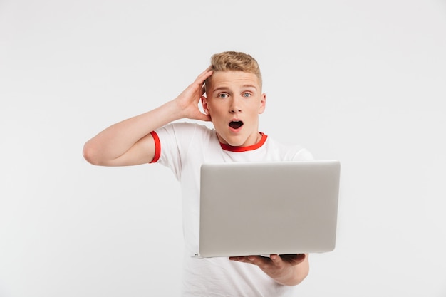 Teenage guy excité 16-18 ans en t-shirt tenant un ordinateur portable ouvert et hurlant de surprise isolated on white