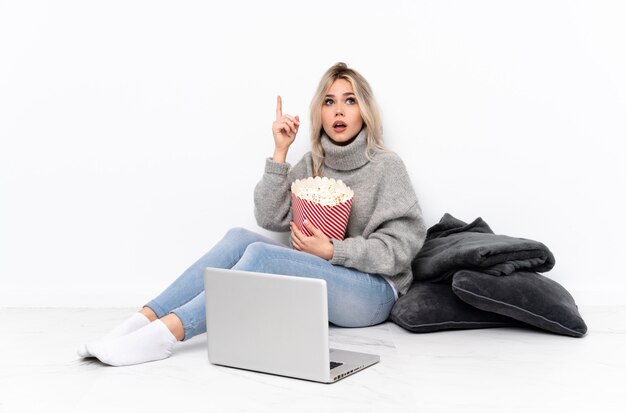 Teenager blonde woman eating popcorn tout en regardant un film sur l'ordinateur portable ayant l'intention de réaliser la solution tout en levant un doigt