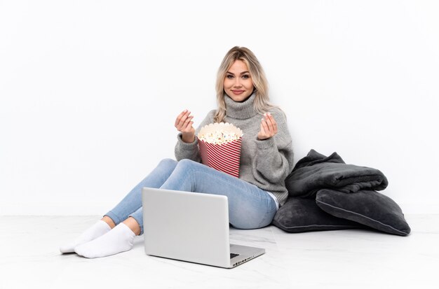 Teenager blonde woman eating popcorn tout en regardant un film sur l'ordinateur portable faisant le geste de l'argent