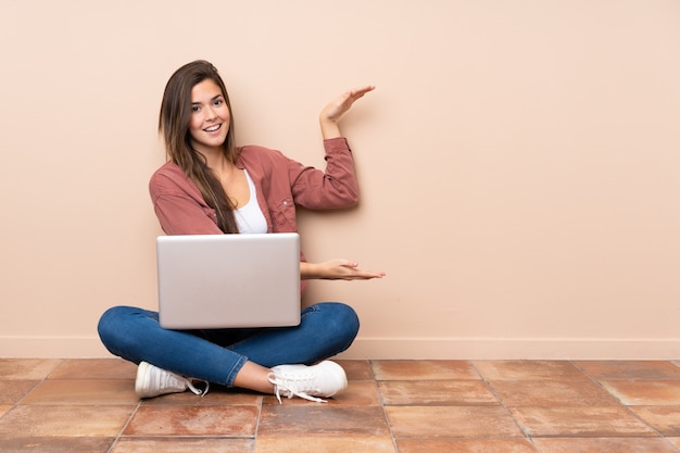 Teenager student girl sitting on the floor avec un ordinateur portable tenant copyspace pour insérer une annonce