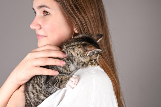 Teengirl tenant un bébé chat sur fond gris mignon souriant tendrement les yeux fermés chaton est assis sur l'épaule de la femme