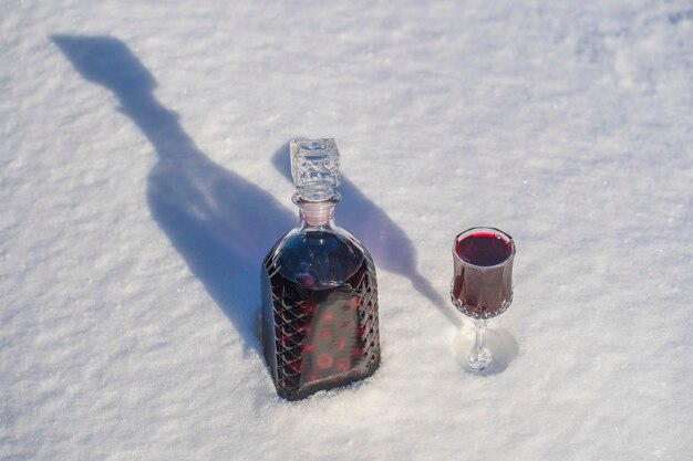 Teinture maison de cerise rouge dans une bouteille en verre et un verre de cristal de vin sur fond blanc et neige