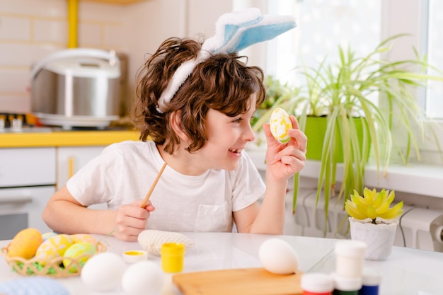 Teinture d'oeufs de pâques garçon bouclé vêtu d'un t-shirt blanc et d'oreilles de lapin bleu peint des oeufs de pâques