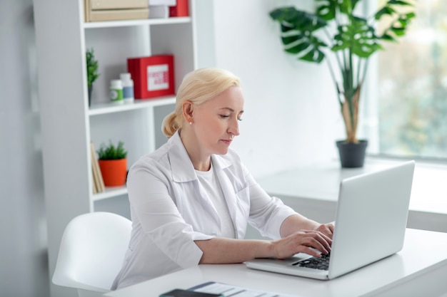 Télémédecine. Praticienne blonde d'âge moyen assise à son bureau et parlant au patient en ligne