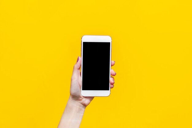 Téléphone blanc avec écran noir dans une main féminine isolée sur un mur jaune de couleur vive